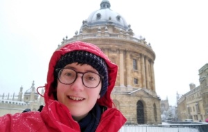 A person smiling wearing a red jacket in front of snow-covered Radcliffe Camera