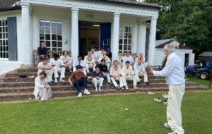 Person talking to a group of people in cricket gear sitting on the steps of a sports pavilion
